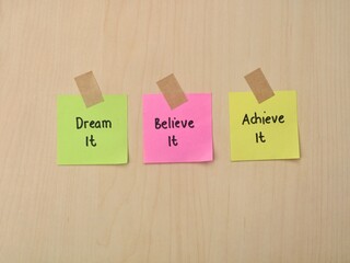 colorful sticky notes with inspirational quotes Dream It, Believe It, Achieve It on a wooden background