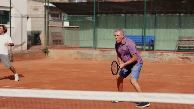 Two players, an adult and an elderly man, play tennis together, they actively move around the court with rackets. Elderly father playing outdoor tennis with his son
