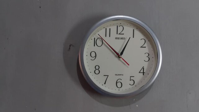Footage of an analog quartz wall clock made in Japan, hanging on a gray painted wall.