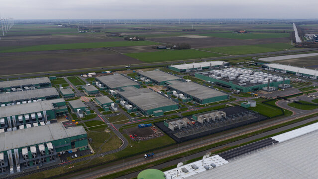 Aerial view of sprawling data centers contrast against the flat landscape, windmills stand distantly on the horizon, Middenmeer, Netherlands.
