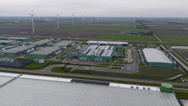 Aerial view of sprawling data centers amidst verdant fields, windmills turning gracefully in the distance, Agriport, Middenmeer, Netherlands.