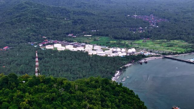 Drone aerial of Pertamina TBBM Manggis, Bali&mdash;coastal fuel terminal with white storage tanks and jetty beside forested hills and quiet bay.