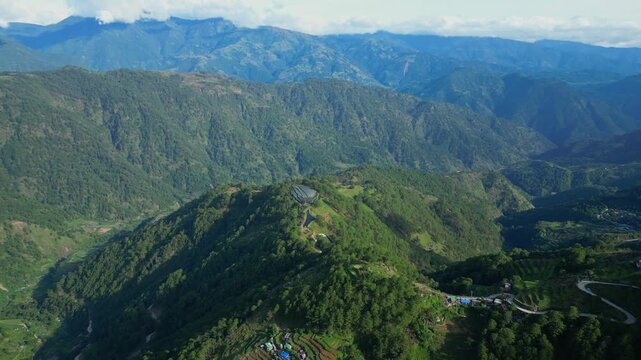 High sweeping aerial turn over a prominent mountain peak in Atok, Benguet, showcasing deep green valleys and distant blue ridges under a clear sky.