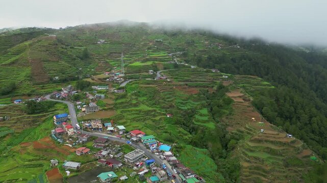 Forward gliding aerial following the winding mountain pass as it cuts through the multi-tiered vegetable terraces and rural clusters of Buguias, Benguet, Philippines.