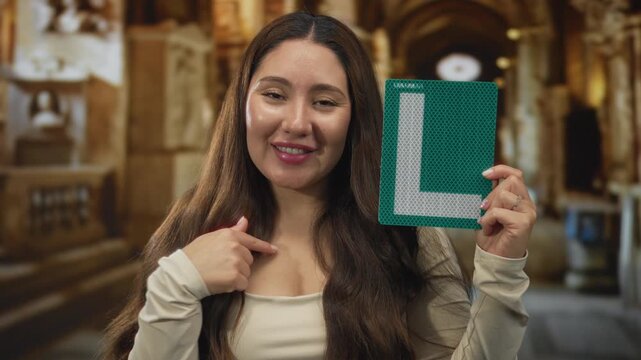 Young hispanic woman holding a green learner sign and pointing to her chest in a building; confidence empowerment pride selfesteem assurance boldness belief courage motivation encouragement.