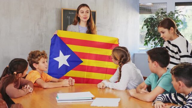 Happy young female teacher holding colorful flag of Estelada while working with preteens schoolgirls and schoolboys in classroom