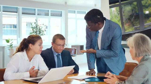 In conference hall, African American man auditor speaks before meeting of shareholders of businessmen. Frustrated, frowning colleagues listen to report on management problems in company
