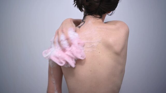 Back view of a young woman scrubbing her skin with a pink loofah sponge and foaming shower gel, engaging in her daily hygiene routine in the bathroom for clean, healthy skin