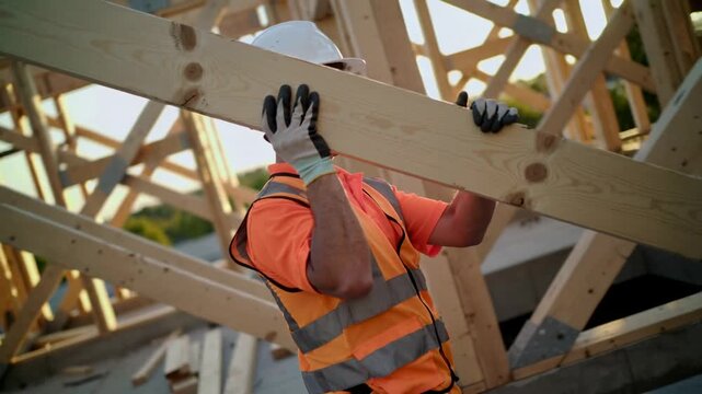Construction worker carrying wooden board at building site wearing helmet, vest, and gloves.
