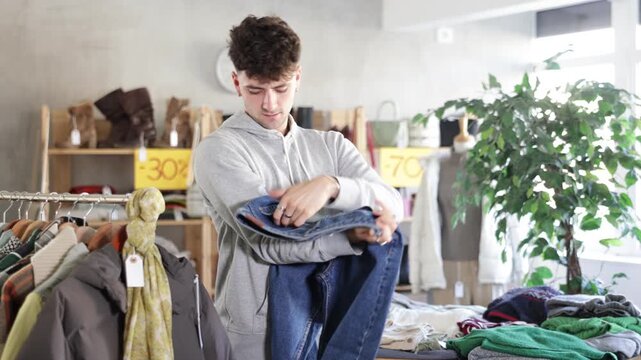 Interested young guy examining stack of jeans during fall sale in clothing store, looking for right fit and style among discounted clothes