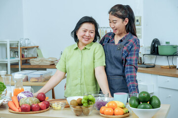 Mature asian mother leaning happily beside adult daughter in bright kitchen with fresh fruits vegetables enjoying family cooking time together creating healthy salad showing love harmony domestic