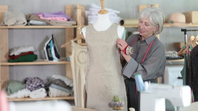 Attentive mature female dressmaker working with tailor's dummy in sewing workshop