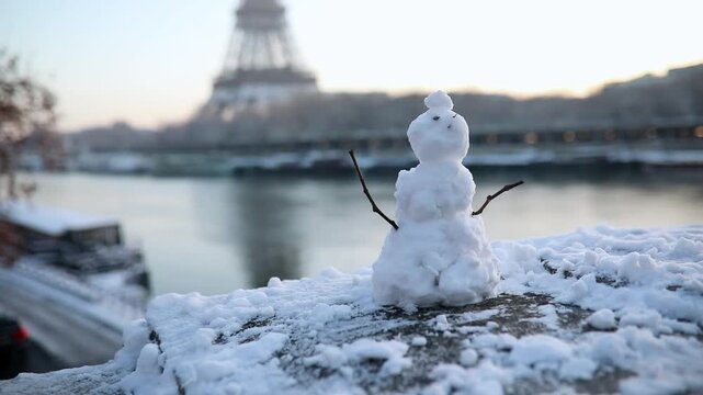 Charming little snowman on a snowy ledge with the Eiffel Tower in the background during a cold winter day in Paris, France. Fun seasonal scene by the Seine River.