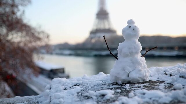 Charming little snowman on a snowy ledge with the Eiffel Tower in the background during a cold winter day in Paris, France. Fun seasonal scene by the Seine River.