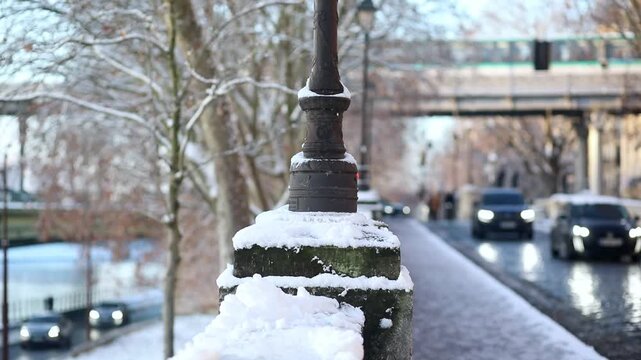 Winter scene in Paris with a close-up of a traditional lamp post covered in snow along a cobblestone street. A car with headlights on is driving in the background