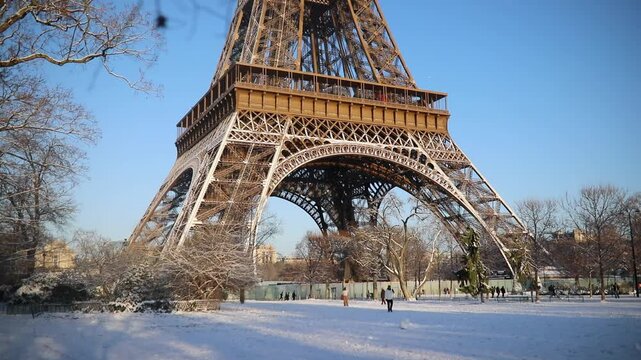 Wide view of the Eiffel Tower with its base surrounded by snow-covered trees and park grounds in Paris. Captured on a bright winter morning, with people walking in the distance and long shadows.