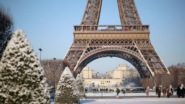 Wide view of the Eiffel Tower with its base surrounded by snow-covered trees and park grounds in Paris. Captured on a bright winter morning, with people walking in the distance and long shadows.