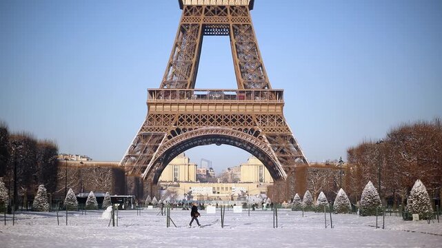 Front view of the Eiffel Tower rising above a snow-covered park in Paris on a sunny winter day. Blue sky, snowy grounds, and trimmed trees create a postcard-perfect scene.