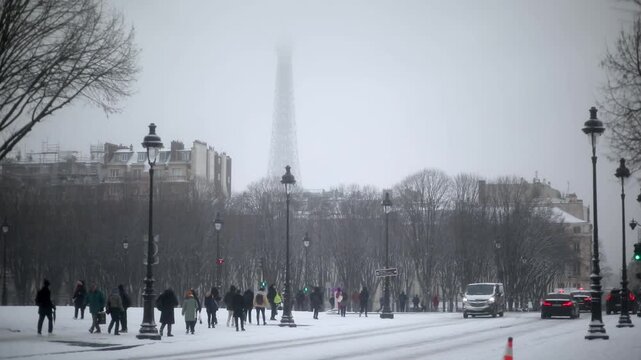 People walk along a snow-covered street in Paris during winter snowfall. The Eiffel Tower is faintly visible through the mist and falling snow.