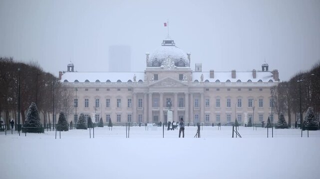 The historic Ecole Militaire building in Paris seen from Champ de Mars during a snowy winter day. Snow blankets the ground and rooftops while the atmosphere is calm and overcast