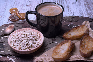 Black mug with coffee foam on tree bark, with a bowl of rolled oats and crispy toasted bread, rustic style
