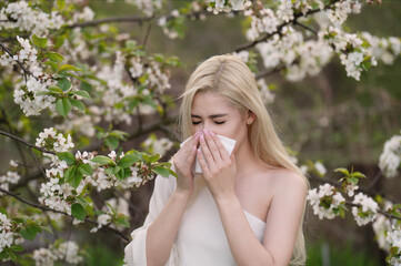 A blonde girl with long hair sneezes and blows her nose into a handkerchief in a blooming garden in spring. Pollen and flowering allergy. Runny nose.