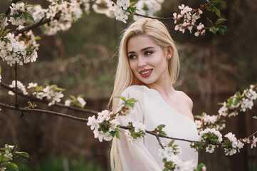 A beautiful girl with long blond hair in a blooming garden, a portrait among branches of apricot and cherry blossoms. Spring is the blossoming of femininity, beauty, and peace.
