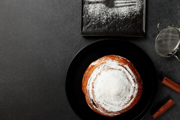 Round, delicious bun with cinnamon and powdered sugar on a black plate on a gray background
