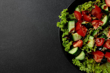 Vegetable salad of tomatoes, cucumbers, onions, lettuce dressed with aromatic dressing in a black plate on a gray background
