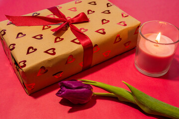 a romantic scene featuring a heart-patterned gift box tied with a red ribbon, accompanied by a lit pink candle and a purple tulip on a vibrant red background.
