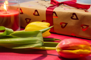 romantic arrangement of two fresh tulips&mdash;red and yellow&mdash;laying in the foreground, with a blurred gift box wrapped in heart-patterned paper and a lit candle in the background.