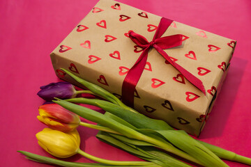 a brown gift box adorned with shiny red hearts and a ribbon, resting beside three colorful tulips on a solid pink background.