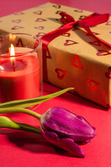 a romantic scene featuring a heart-patterned gift box tied with a red ribbon, accompanied by a lit pink candle and a purple tulip on a vibrant red background.