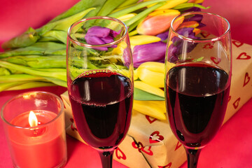 a romantic celebration featuring two champagne flutes filled with red wine, a heart-patterned gift box, fresh tulips, and a lit candle on a red background.