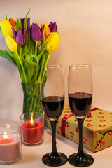 a romantic setting featuring two glasses of red wine, a heart-patterned gift box, and a vase of colorful tulips illuminated by a lit red candle.
