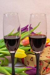 two glasses filled with red wine standing in front of a heart-patterned gift box and a colorful bouquet of fresh tulips.