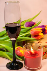 a single glass of red wine standing beside a lit candle, with a bouquet of colorful tulips and a wrapped gift box in the soft background.