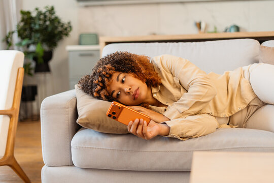 Young woman feeling bored on couch, scrolling smartphone at home