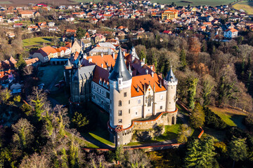 Zleby Castle is shown from the air, surrounded by trees and a small village. The castle has distinctive towers and a red roof. It is located in the countryside.