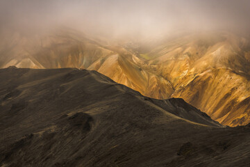 View of stark black ridges contrast with the warm, ochre hues of distant peaks veiled in mist, creating a surreal, ethereal landscape, Landmannalaugar, Central Highland, Iceland.