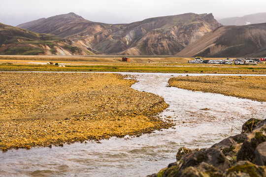 View of a winding river cuts through a golden gravel landscape, leading the eye toward distant, rugged mountains under a cloudy sky, Landmannalaugar, Iceland.