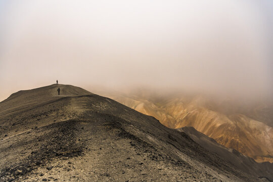 View of two distant figures trek along a stark, grey mountain ridge, where mist veils the colorful slopes below, creating an ethereal atmosphere, Landmannalaugar, Iceland.