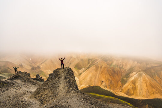View of figures triumphantly poised atop a rugged peak amidst a surreal, mist-shrouded landscape of ochre and gray mountains, Landmannalaugar, Central Highland, Iceland.
