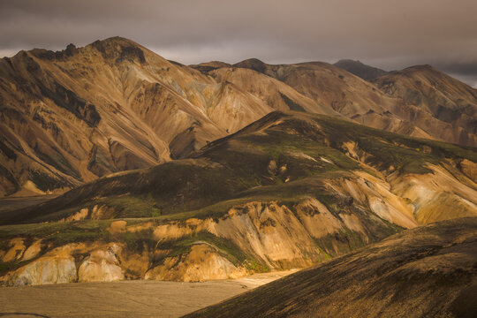View of rugged, golden mountains rise under a brooding sky, their slopes patched with green, creating a stark contrast of color and texture, Landmannalaugar, Iceland.