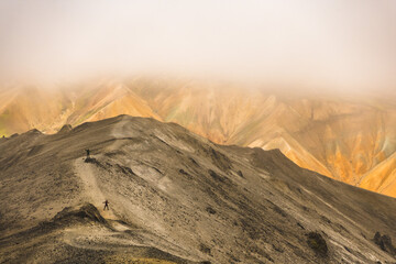 View of hikers ascending a stark grey ridge towards vibrant, ochre mountains veiled in mist, a journey through Iceland's raw, elemental beauty, Landmannalaugar, Iceland.
