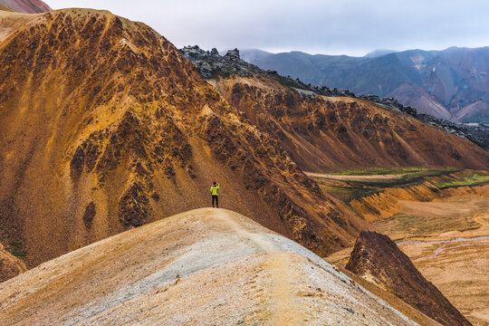 View of a lone figure stands atop a ridge, overlooking a landscape of ochre and grey mountains under a somber sky, Landmannalaugar, Central Highland, Iceland.