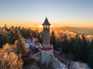 Stepanka lookout tower stands tall in the Jizera Mountains during winter. Visitors enjoy a sunset view over the landscape and surrounding trees in this serene mountain setting.
