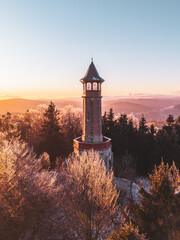 Stepanka lookout tower stands tall in the Jizera Mountains during winter. Frost covers the trees as the sun sets, casting warm light on the landscape. A peaceful scene invites exploration.