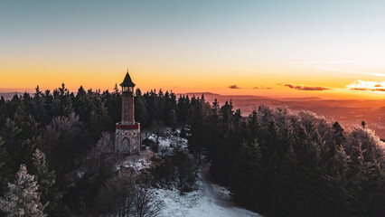 The Stepanka lookout tower stands in the Jizera Mountains during winter. Snow covers the ground while the sunset casts warm colors over the landscape. Trees surround the tower.