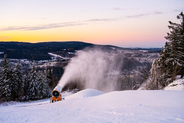 A snow cannon produces snow on a ski slope at sunset. Snow covers the ground, and mountains stretch in the background. Skiers will enjoy this area for winter sports soon.